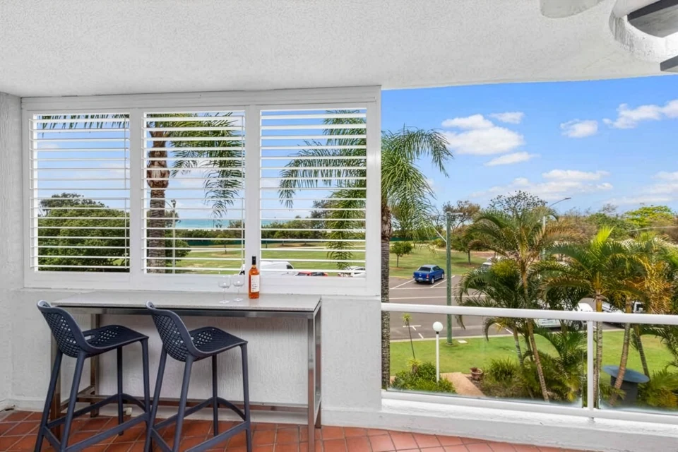 A balcony at Beach Health Retreat with two stools at a high bench, looking through white shutters over palms and parkland to the sea.