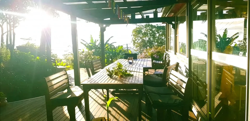 The sun deck of the Bellbunya community house at sunset, a long timber table and chairs beneath bunting, with gardens and trees beyond.