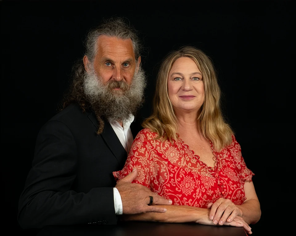 Deb and Ian together against a dark backdrop, forearms resting on a table, both looking towards the camera.