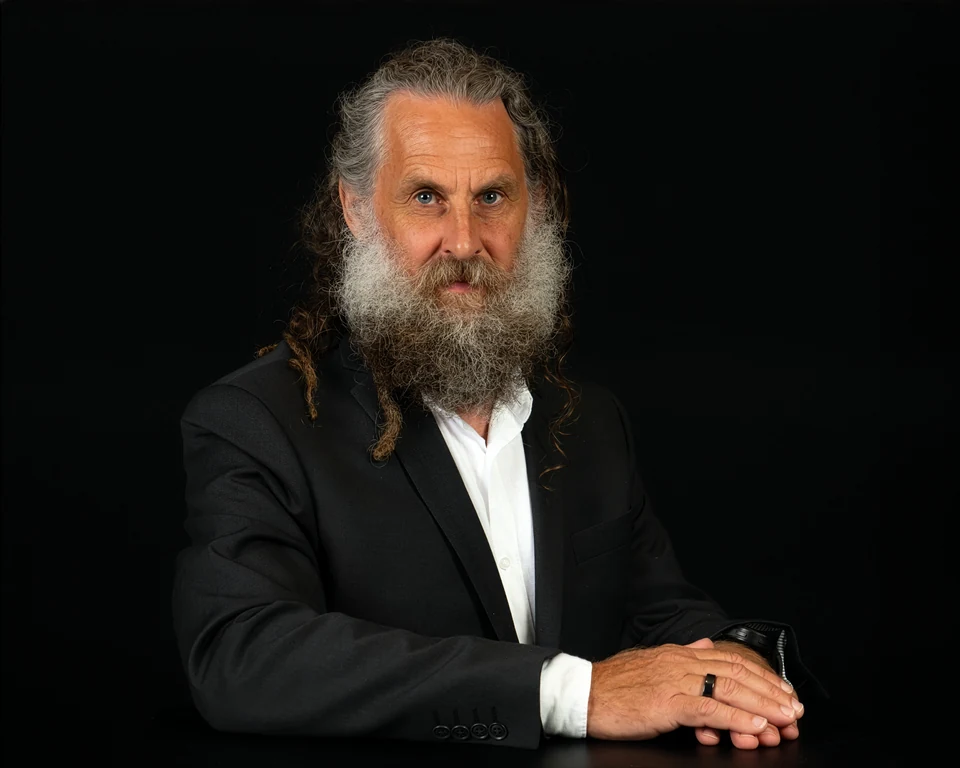 Ian seated at a dark table in a black jacket and open-collared white shirt, looking towards the camera.